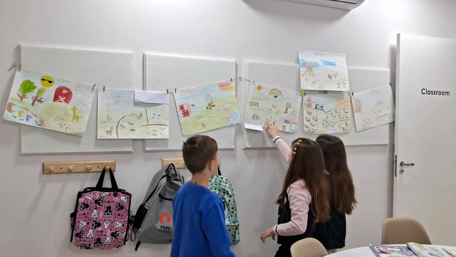 Crianças vistas de costas compartilham desenhos expostos na parede da sala